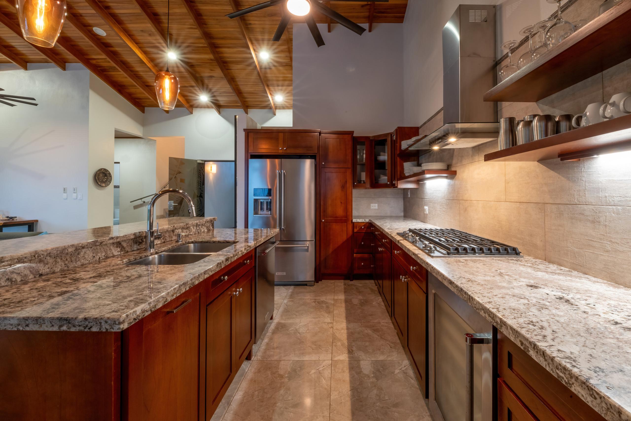 A well-appointed kitchen features rich wood cabinetry, speckled granite countertops, stainless steel appliances, and a vaulted ceiling with exposed wooden beams and ambient lighting.
