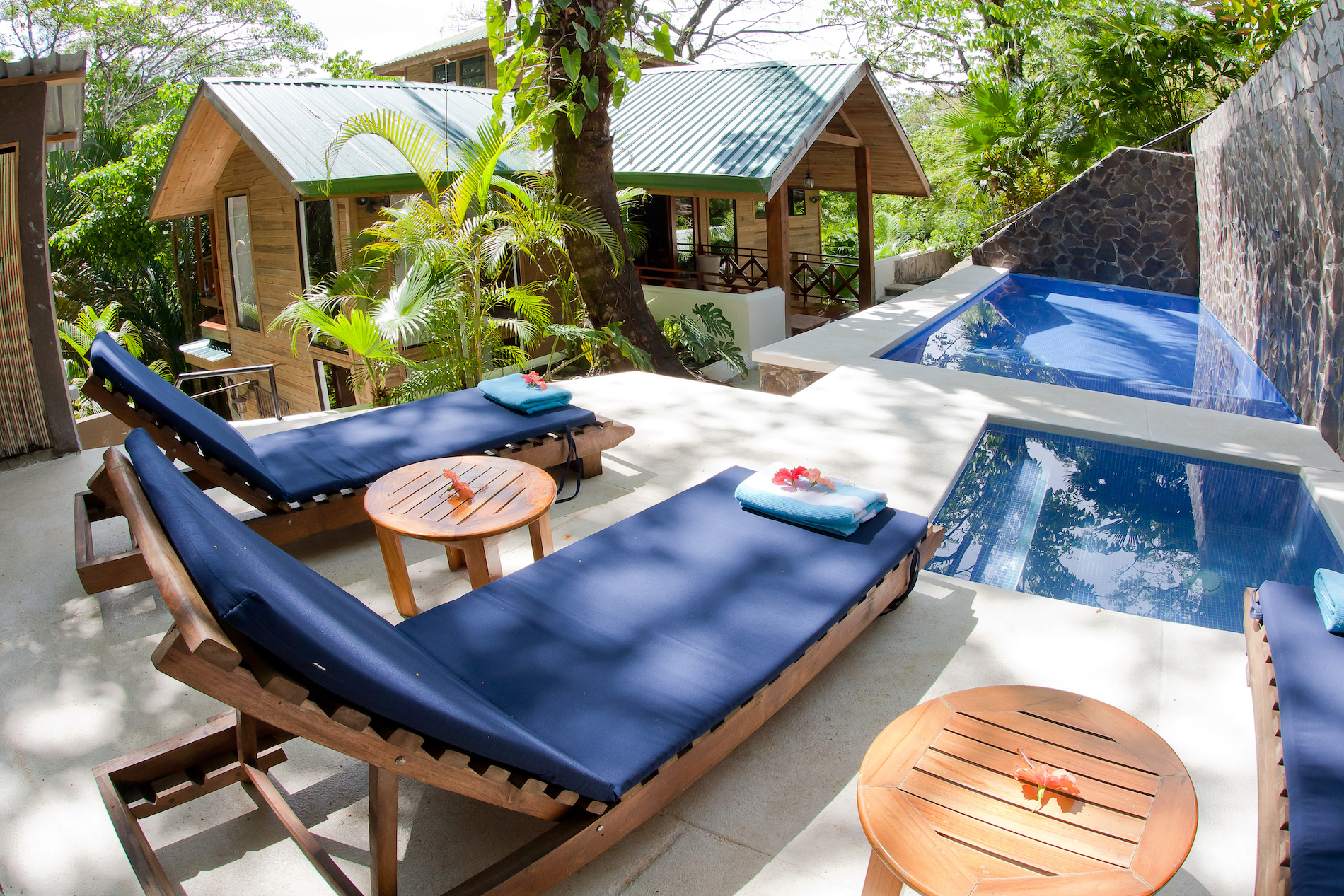 Una terraza con tumbonas y una piscina privada y jacuzzi ofrece vistas a la villa de madera y la exuberante selva tropical.