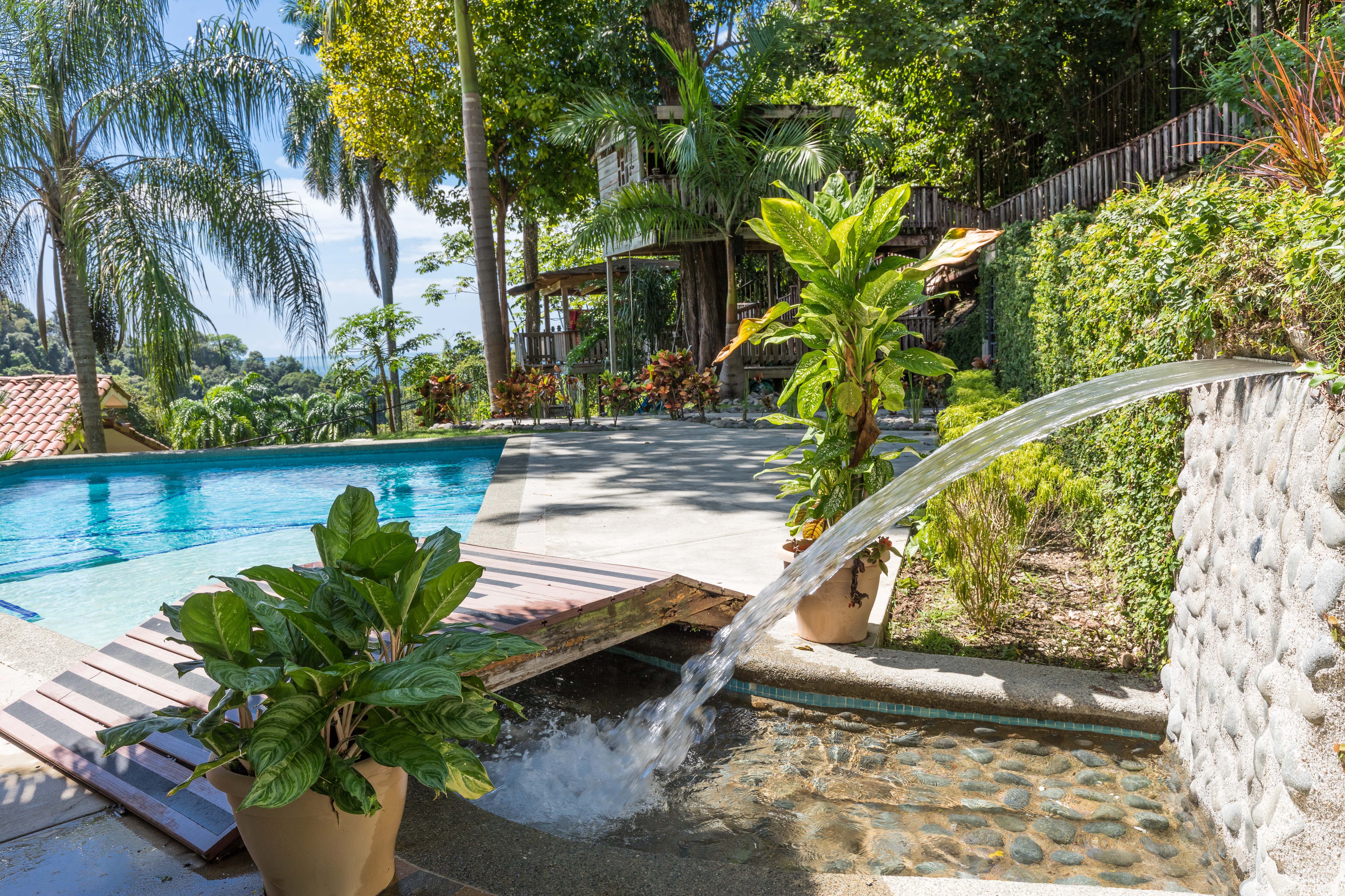 A sunlit blue swimming pool features a cascading water element, surrounded by lush tropical greenery and a wooden treehouse structure.