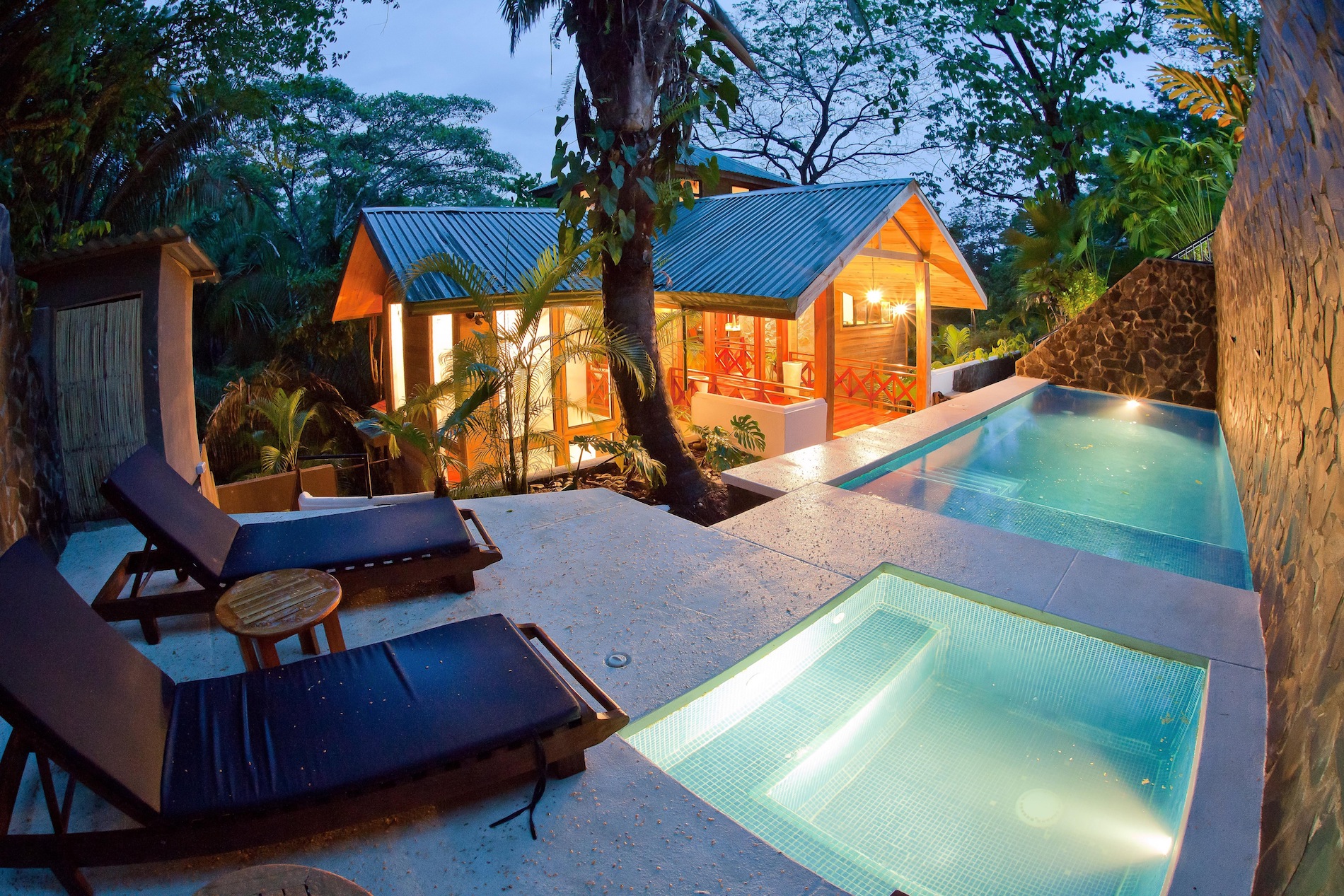 Una piscina privada iluminada y jacuzzi separado se sientan en una terraza de piedra con vistas a una villa de madera de estilo cálido y exuberante follaje en la selva.