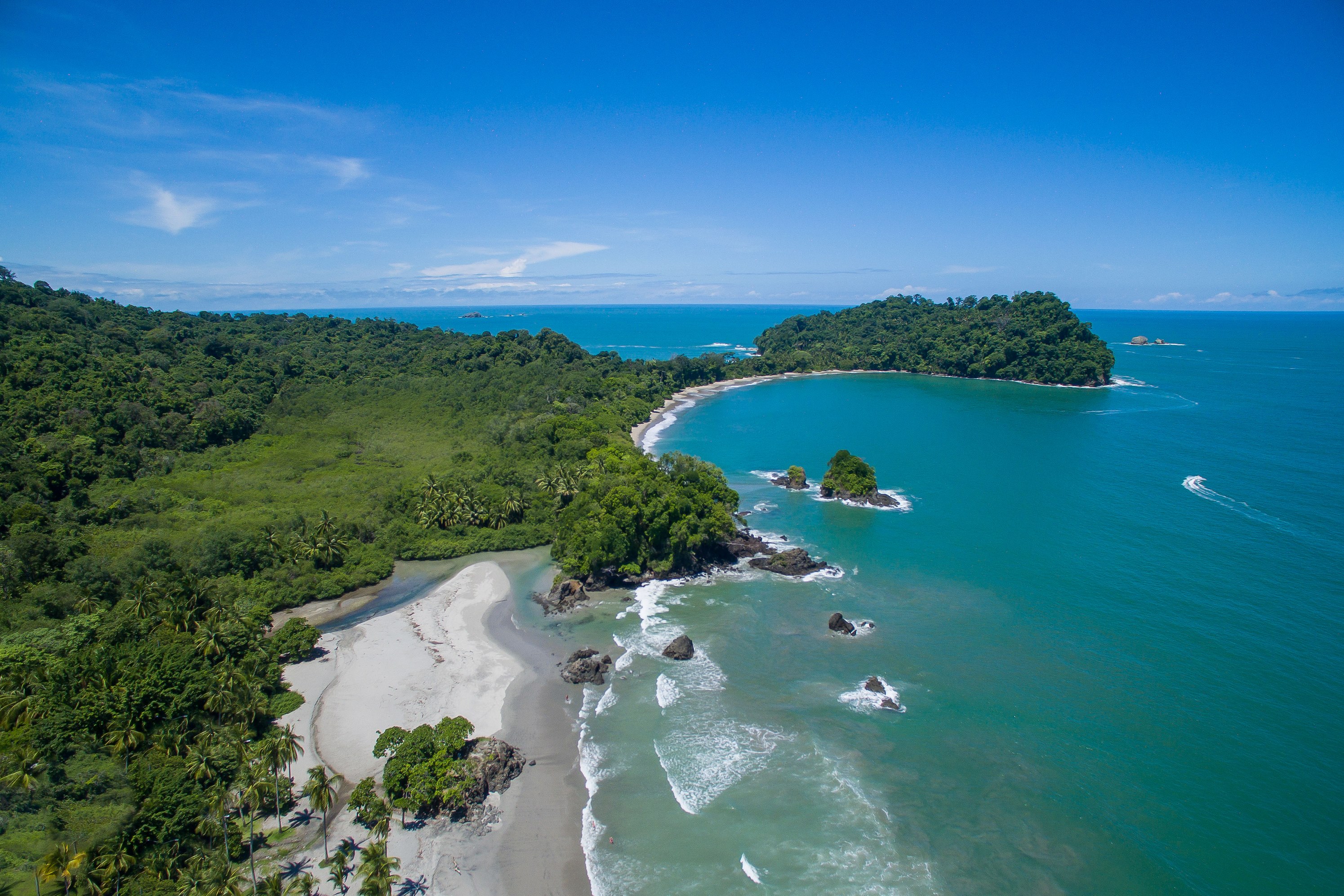 An aerial perspective reveals a tropical coastline with lush rainforest bordering a white sand beach and clear turquoise ocean featuring several rocky islets.