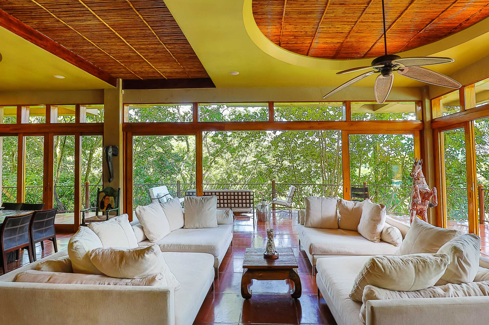 The living room features multiple large sofas, a natural wood paneled ceiling, and sliding windows walls offering direct views of the tropical rainforest and plentiful natural light.