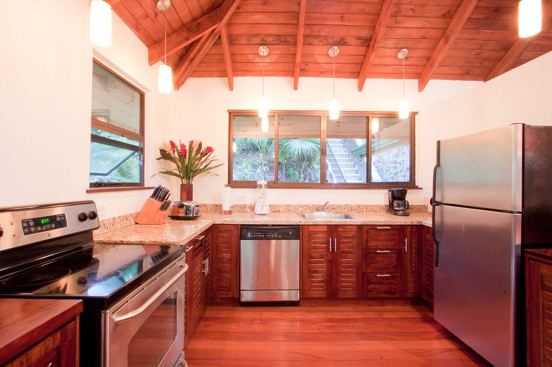 Una cocina bien equipada cuenta con suelos de madera, armarios y techo abovedado, complementado con electrodomésticos de acero inoxidable y ventanas con vistas a la selva tropical.