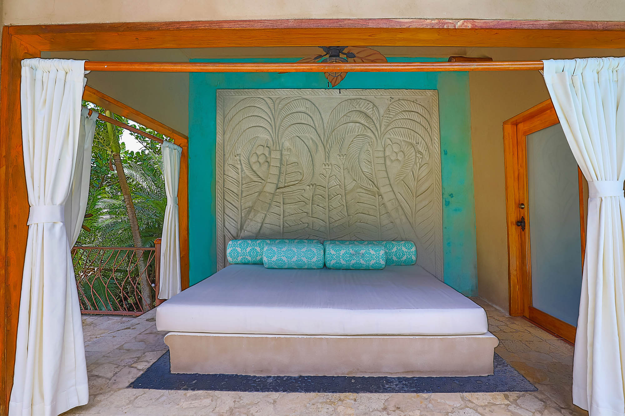 An open-air day bed with a carved headboard and natural wood accents, overlooking a lush tropical rainforest and direct pool access.