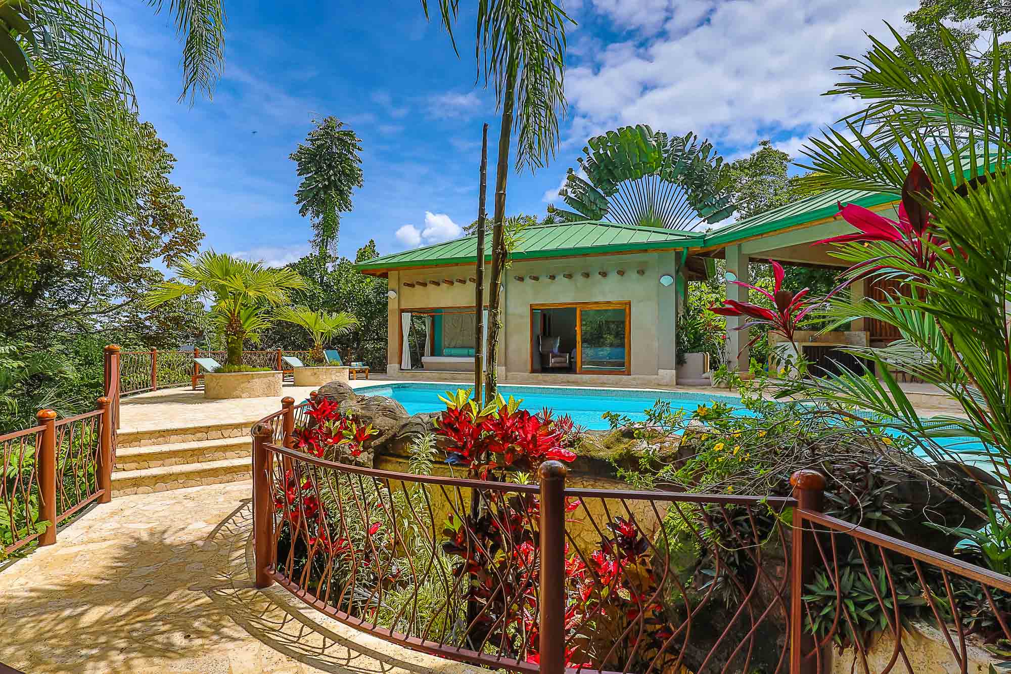 A vibrant blue swimming pool sits amidst abundant tropical plants and palm trees, opening to the natural surroundings.