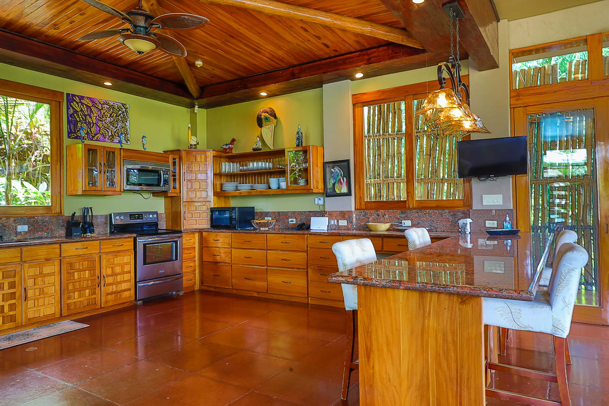 The kitchen, features extensive wooden cabinetry, granite countertops, and a large island, providing tropical views through wooden-framed windows. 