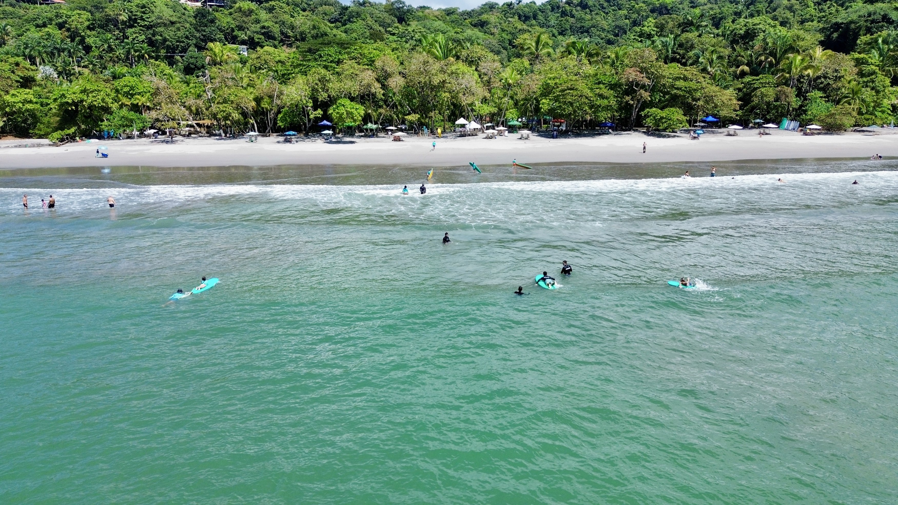 An aerial view shows a sun-drenched sandy beach with people swimming and surfing in the turquoise ocean, backed by a lush green jungle-covered hillside.