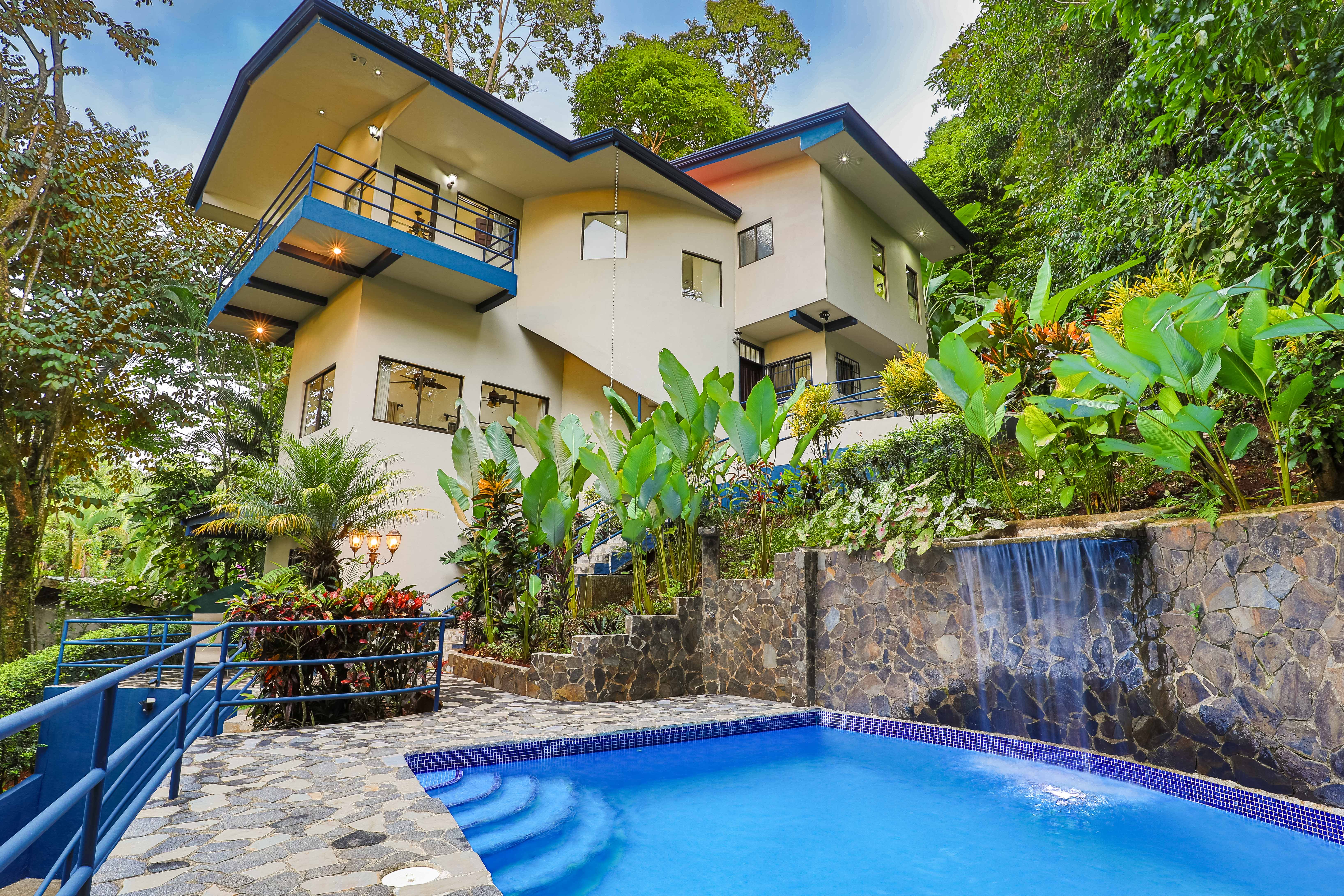 The multi-level contemporary villa with blue trim overlooks a private pool featuring a waterfall, surrounded by dense rainforest vegetation.