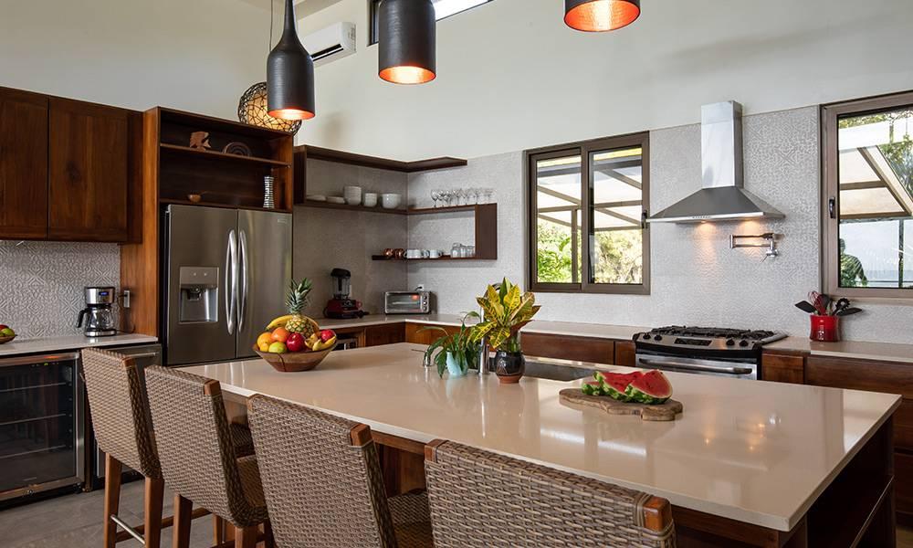 Esta cocina bien iluminada cuenta con armarios de madera oscura, una amplia isla central con asientos de barras, electrodomésticos de acero inoxidable y grandes ventanas que ofrecen vistas al follaje tropical circundante.