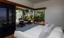 Un luminoso dormitorio con techo abovedado cuenta con grandes puertas de vidrio que se abren a un patio privado con sillas de salón, jardín tropical y una vista parcial al mar.