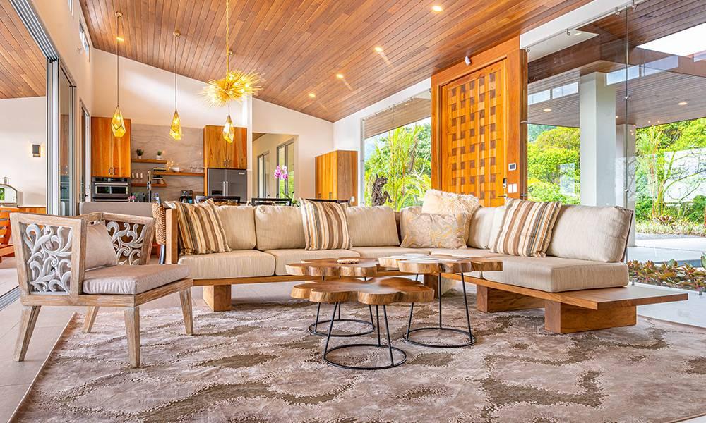 Una sala de estar abierta con un techo inclinado de madera y un amplio sofá seccional se conecta a una cocina adyacente, con paredes de vidrio de altura completa que ofrecen vistas a la selva circundante.