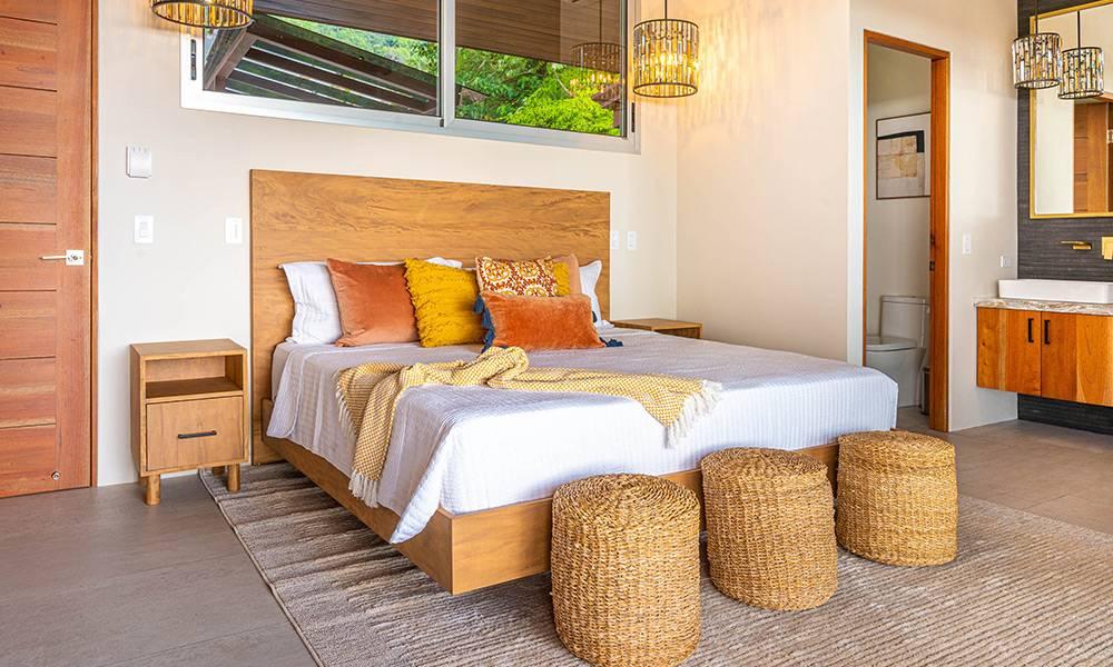 A bright bedroom features a king-size bed with a wooden headboard, an en-suite bathroom visible through an open doorway, and a large window offering jungle views.