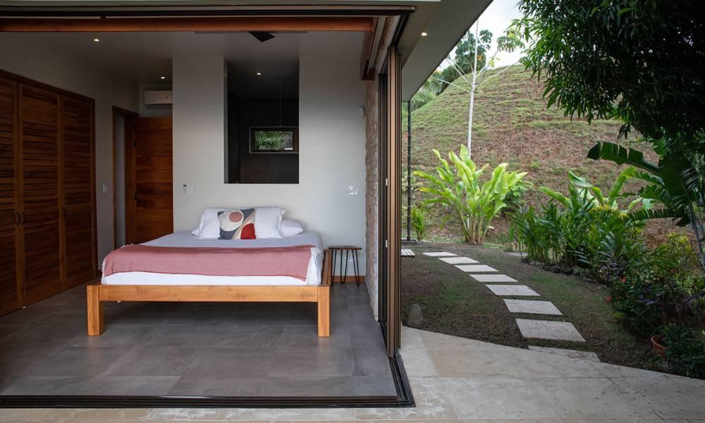 Un dormitorio con cama king cuenta con puertas dobles que se abren directamente a una vía de jardín y ladera exuberante, proporcionando luz natural y una conexión al aire libre.