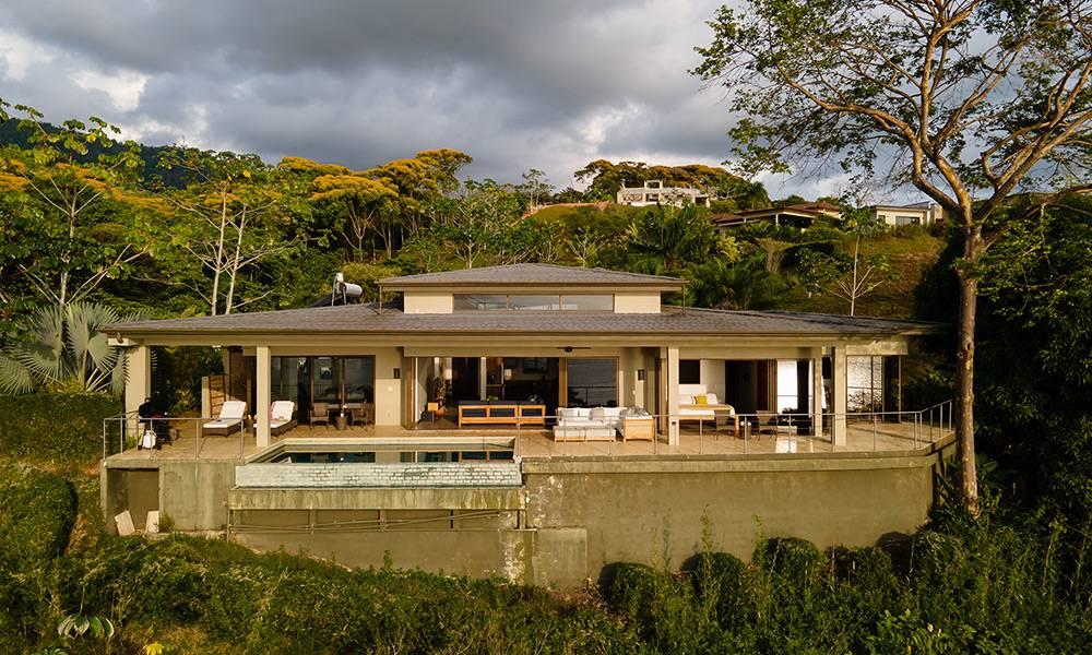 La villa de un solo nivel cuenta con una amplia terraza cubierta con una piscina visible, al aire libre y amplias puertas de vidrio que se abren a un exuberante entorno tropical.