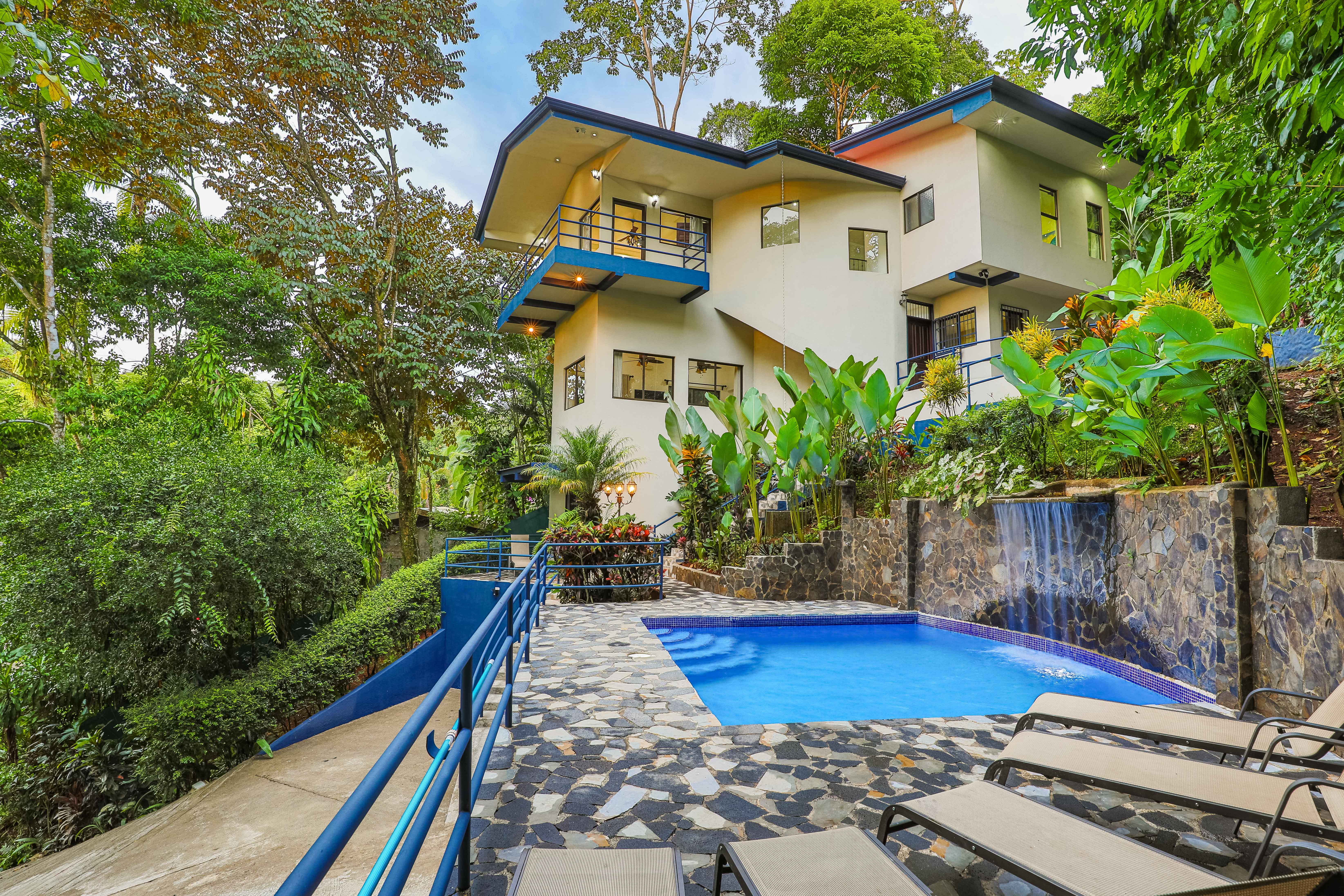 Una villa multinivel contemporánea con acentos azules, múltiples balcones y grandes ventanales con vistas a una piscina privada con cascada, todo situado dentro de una vibrante selva tropical.